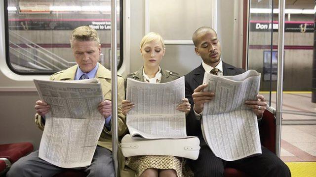 Two men and woman sitting in subway train side by side, reading newspaper